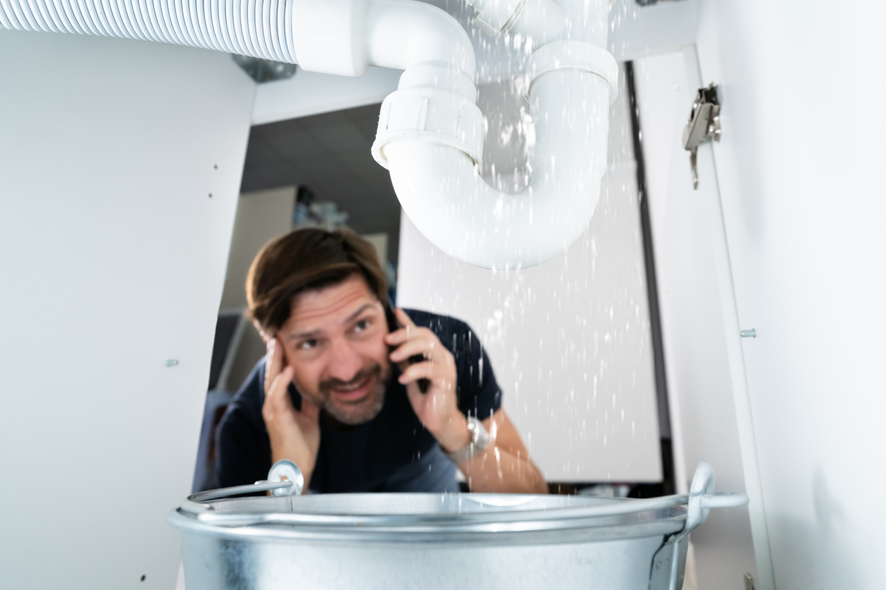 Worried Man Calling Plumber While Watching Water Leaking From Sink